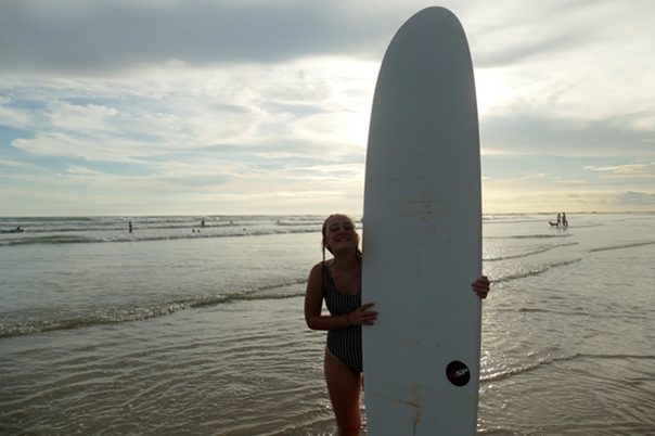 A girl is smiling and standing behind a surf board on a beach.