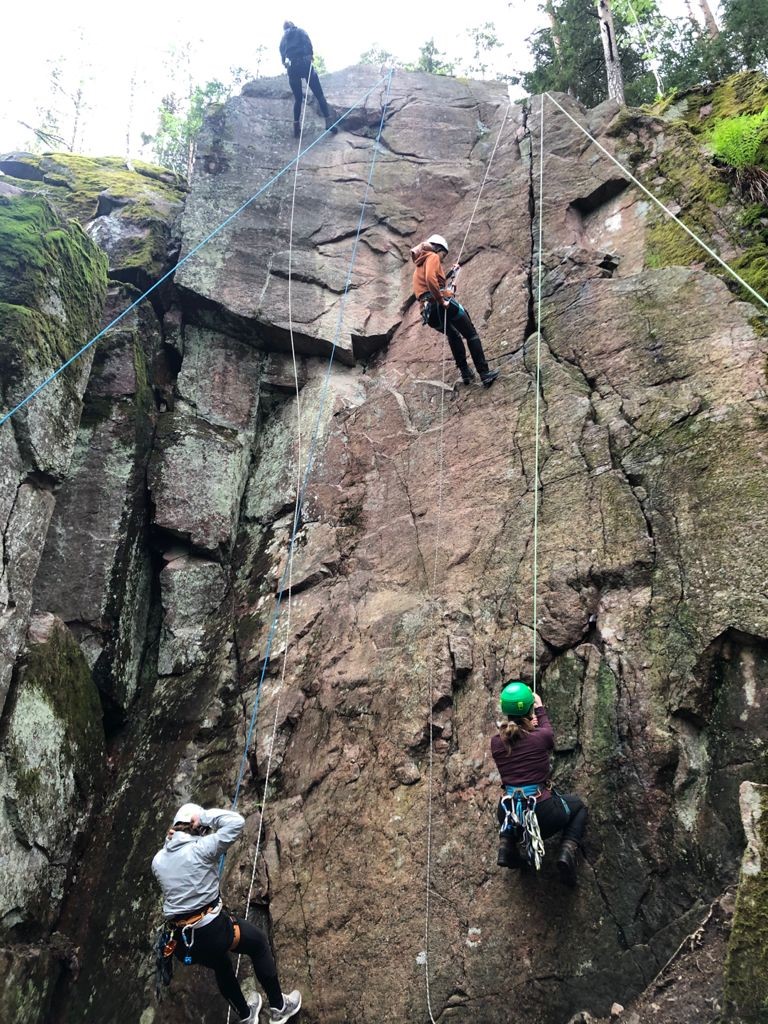 Four students in protective gear are climbing up a very steep rock in a forest setting.