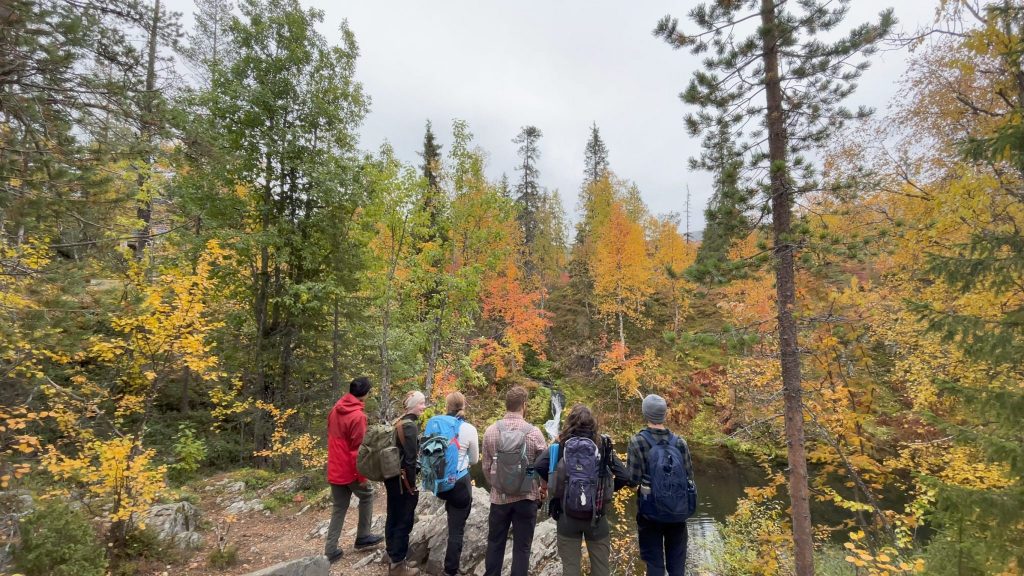 Six students standing on the edge of a cliff looking over a mountain pond surrounded by trees in full autumn foliage.