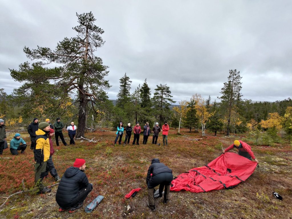 A group of students in outdoor gear gathered around their instructors who are teaching how to set up a tent. Surrounded by autumnal Lapland scenery.