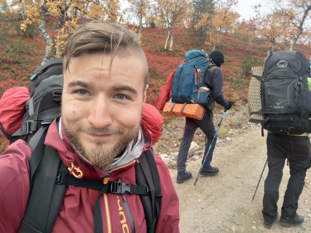 A selfie of a smiling student with two other students in the background. They are trekking/hiking in nature.